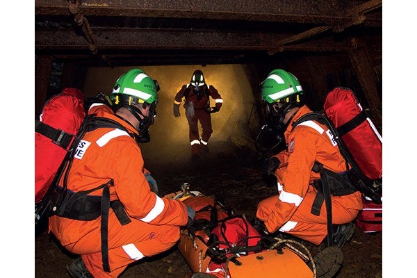 Three men in a confined space with gas., wearing orange overalls and green helmets, carrying  safety equipment and saving a man who’s on the floor in recovery apparatus