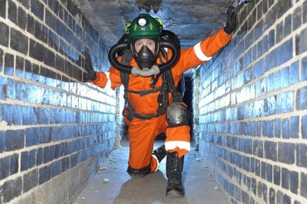 Man wearing orange overalls kneeling down in a confined space wearing breathing apparatus 