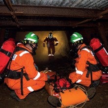 Three men in a confined space with gas., wearing orange overalls and green helmets, carrying  safety equipment and saving a man who’s on the floor in recovery apparatus